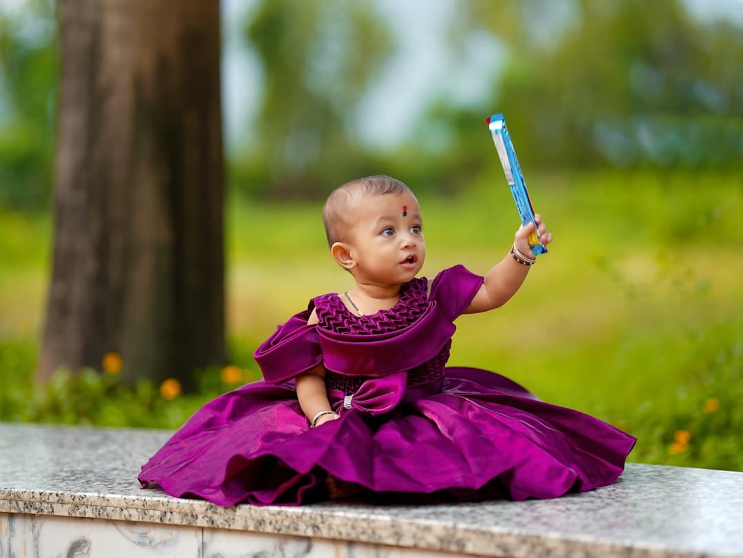 A baby in a purple dress holds a blue toy.